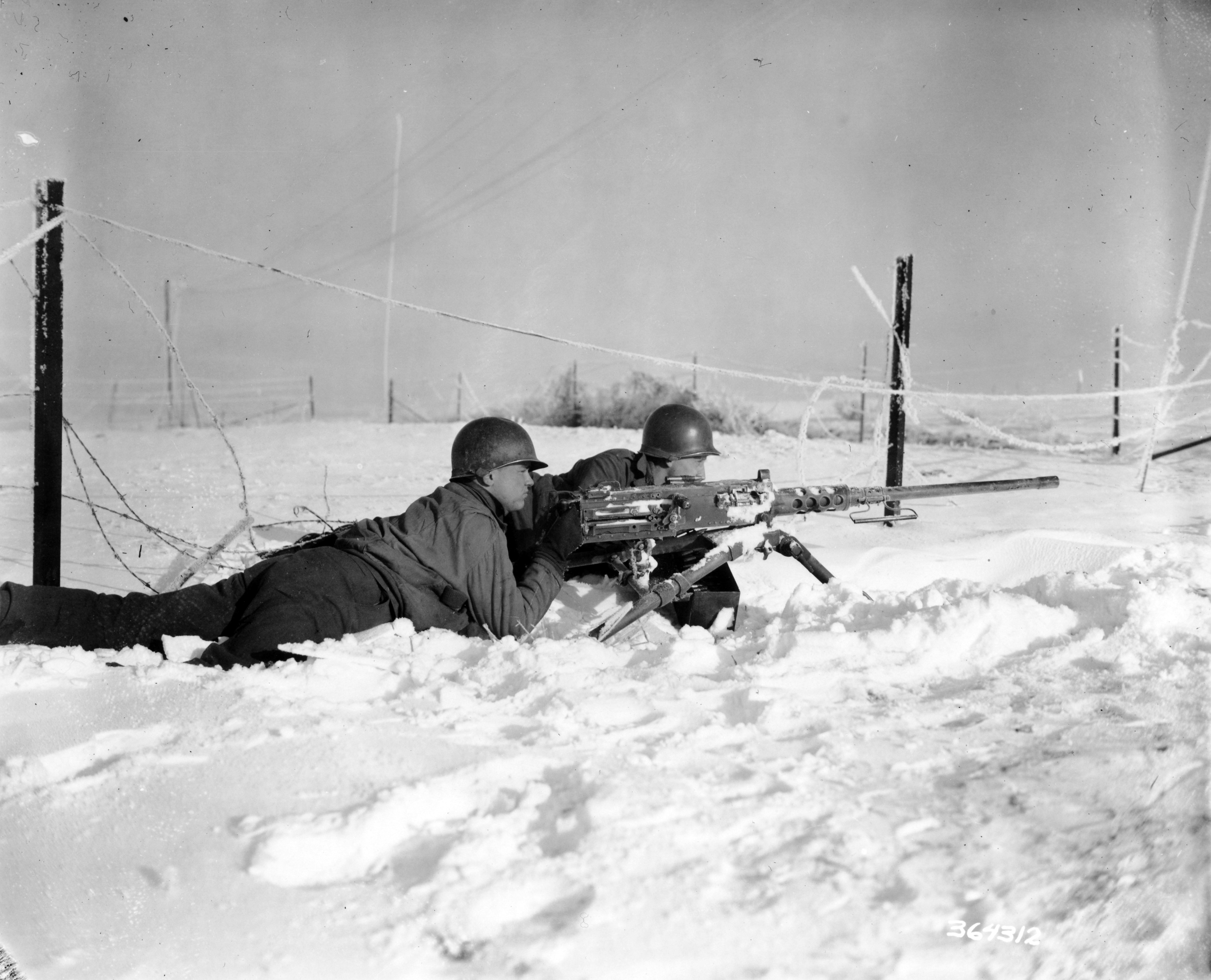 Gunner Pvt. Nick Hotolsky, Russellton, Pa. and gunner Pfc. Roman Vinicki, South Bend, Ind., laying in the snow and guarding a road junction with their .50 caliber machine gun.